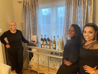 three women standing in front of a living room with bottles of wine